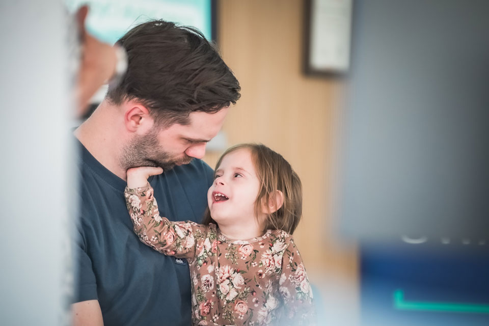 A young girl smiles up at an adult man holding her, with a soft blurred background creating a warm and intimate moment.
